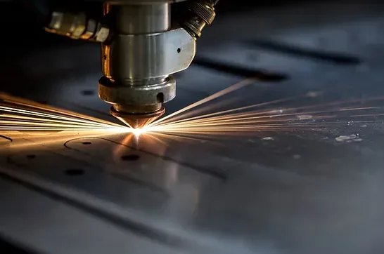 A CNC laser cutting head slicing a flat metal sheet with a fan of bright sparks on a fabrication floor