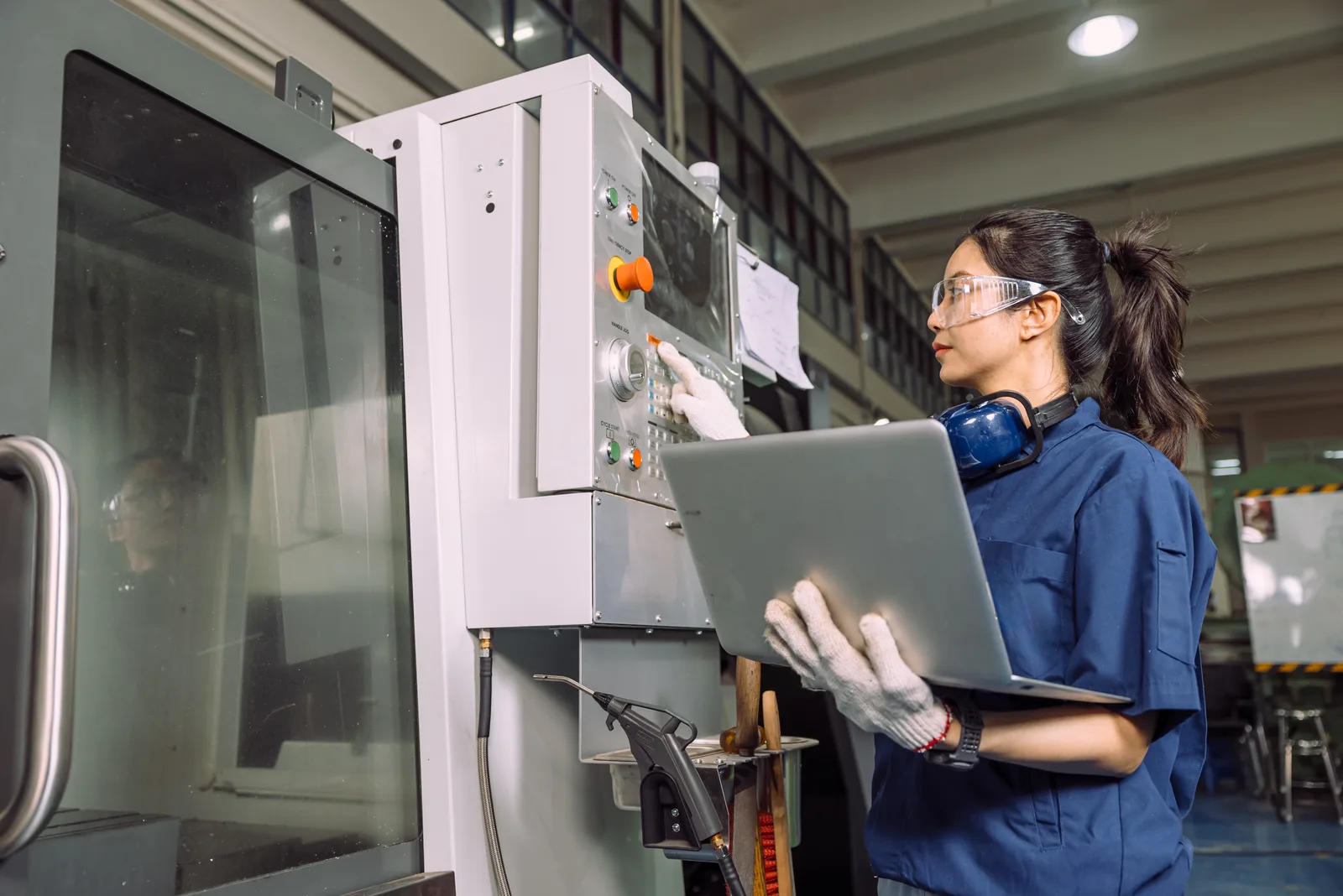 A machinist in safety glasses and ear defenders working a machine control panel while holding a laptop